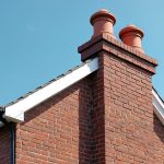 Red brick house gable with chimney stacks against clear blue sky. White fascia and black guttering, visible on roof edge. Architectural detail.